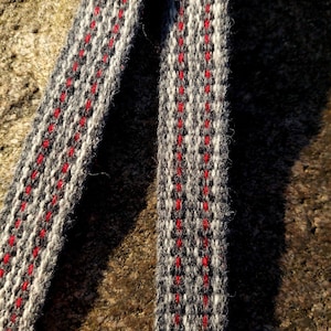 May include: Close-up of two woven straps with a repeating pattern. The straps are primarily gray with white and red accents. The texture of the weave is visible, and the straps appear to be made of a soft material. The background is a rough, textured surface.