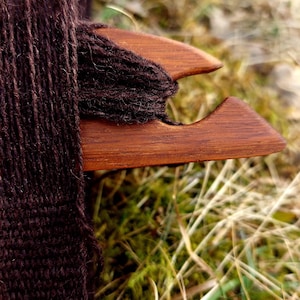 Peut inclure: Une navette en bois avec du fil brun foncé enroulé autour. La navette est d'un riche brun rougeâtre, et le fil est d'un brun profond et texturé. L'arrière-plan présente de l'herbe verte et un feuillage brun doré sec.