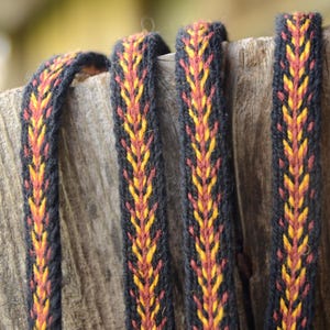 Peut inclure: Gros plan sur quatre lanières tissées avec un fond noir et un motif central de chevrons orange et jaunes. Les lanières sont drapées sur une surface en bois vieilli, mettant en valeur la texture et la couleur du matériau.
