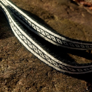 May include: Close-up of two black and white woven straps with a repeating geometric pattern. The straps are lying on a rough, brown surface. The design features a series of white diamond shapes against a black background.