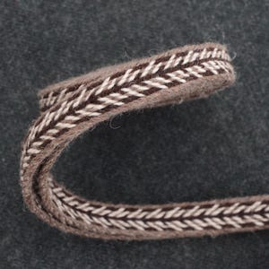 May include: Close-up of a curved piece of brown and white woven trim. The trim has a textured appearance with a repeating chevron pattern in alternating brown and white colors. The background is a dark gray fabric.
