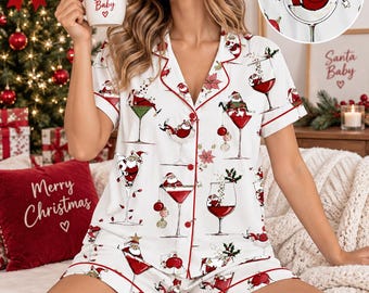 May include: A white short-sleeve pajama set with red piping, featuring a repeating print of Santa Claus relaxing in various cocktail glasses, accented with poinsettias and Christmas ornaments. The shirt has a button-down front and a notched collar.