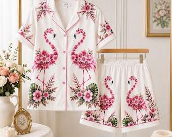 Könnte beinhalten: Ein weißes Pyjama-Set bestehend aus einem kurzärmeligen Hemd mit Knopfleiste und passender Shorts. Der Stoff zeigt ein wiederkehrendes Muster aus rosa Flamingos, die aus floralen Elementen bestehen, akzentuiert durch grüne Blätter und rosa Paspeln.