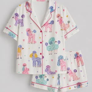 K&ouml;nnte beinhalten: Ein wei&szlig;es Pyjama-Set bestehend aus einem kurz&auml;rmeligen Hemd mit Knopfleiste und passender Shorts. Der Stoff zeigt ein wiederkehrendes Muster aus bunten Pudeln in Rosa, Lila, Gelb und T&uuml;rkis, akzentuiert durch kleine Herzen und Punkte.