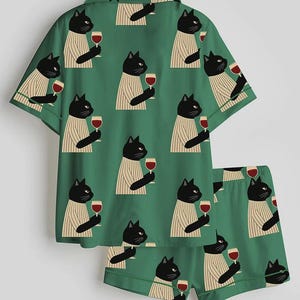 K&ouml;nnte beinhalten: Gr&uuml;nes Pyjama-Set mit einem sich wiederholenden Muster aus schwarzen Katzen in gestreiften Pullovern, die Weingl&auml;ser halten. Das Set besteht aus einem kurz&auml;rmeligen Hemd und passenden Shorts.