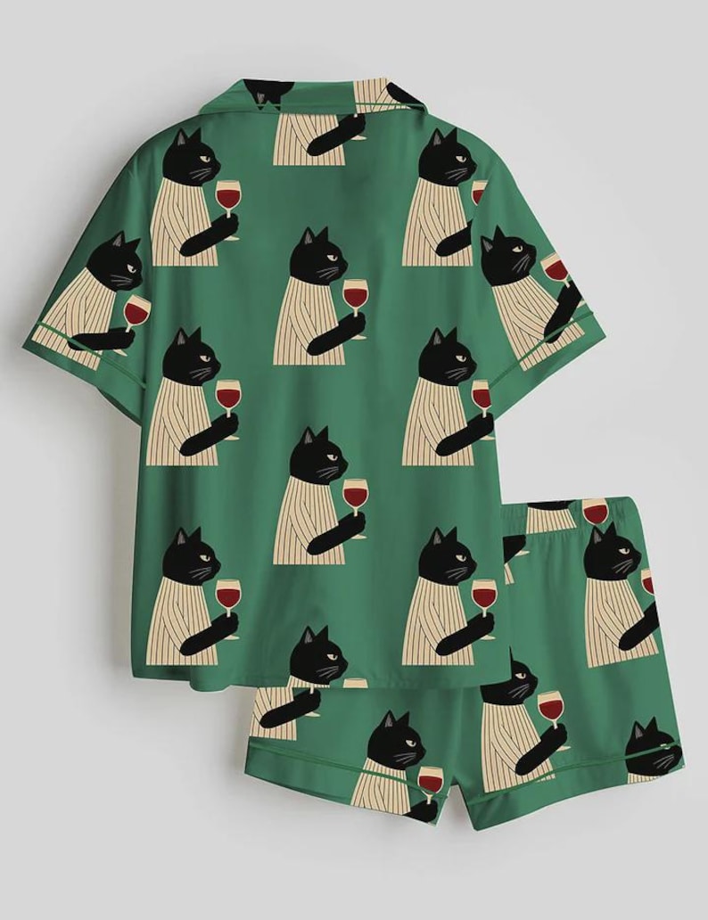 K&ouml;nnte beinhalten: Gr&uuml;nes Pyjama-Set mit einem sich wiederholenden Muster aus schwarzen Katzen in gestreiften Pullovern, die Weingl&auml;ser halten. Das Set besteht aus einem kurz&auml;rmeligen Hemd und passenden Shorts.