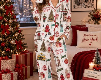 May include: A white long-sleeve button-down pajama set featuring a repeating print of New York City landmarks, Christmas trees, and shopping motifs with red piping. The mug held in the hand displays the text Christmas in New York.