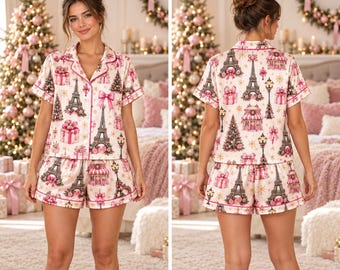 May include: A two-piece pajama set featuring a short-sleeve button-down shirt and matching shorts. The fabric is cream-colored with a repeating print of the Eiffel Tower, Christmas trees, gift boxes, street lamps, and pink bows.