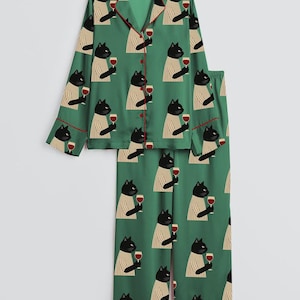 K&ouml;nnte beinhalten: Gr&uuml;nes Pyjama-Set mit einem sich wiederholenden Muster aus schwarzen Katzen, die Weingl&auml;ser halten. Die Pyjamas haben rote Paspeln und ein Oberteil mit Kn&ouml;pfen und langen &Auml;rmeln. Die Hose ist gerade geschnitten.