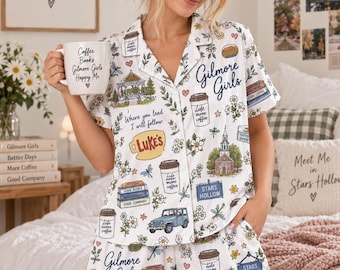 May include: A white pajama set featuring a short-sleeved button-down shirt and matching shorts with a colorful print of Gilmore Girls icons, including coffee cups, books, a gazebo, a church, a blue jeep, and the text Luke's and Stars Hollow.