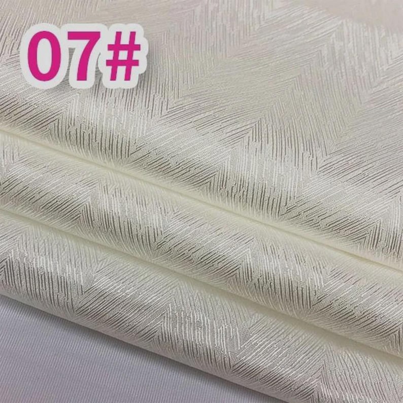 K&ouml;nnte beinhalten: Off-white Stoff mit einem dezenten, strukturierten Fischgr&auml;tmuster. Der Stoff ist gefaltet und zeigt mehrere Schichten. Ein rosa Aufkleber in der oberen linken Ecke zeigt die Nummer "07#".