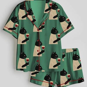 K&ouml;nnte beinhalten: Gr&uuml;nes Pyjama-Set mit einem sich wiederholenden Muster aus schwarzen Katzen, die Weingl&auml;ser halten. Das kurz&auml;rmelige Oberteil und die Shorts haben rote Paspeln. Die Katzen tragen gestreifte Pullover. Das Set ist aus weichem, leichtem Stoff.