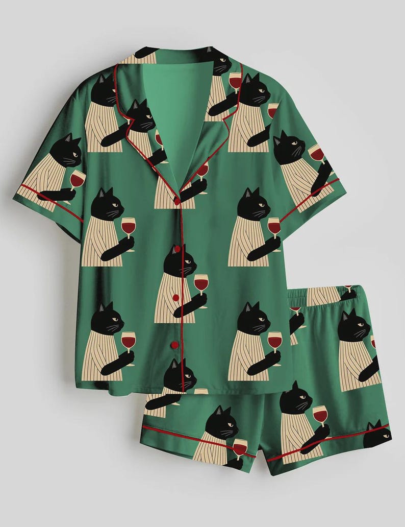 K&ouml;nnte beinhalten: Gr&uuml;nes Pyjama-Set mit einem sich wiederholenden Muster aus schwarzen Katzen, die Weingl&auml;ser halten. Das kurz&auml;rmelige Oberteil und die Shorts haben rote Paspeln. Die Katzen tragen gestreifte Pullover. Das Set ist aus weichem, leichtem Stoff.