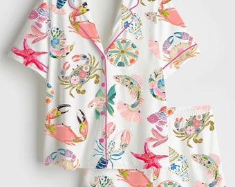 May include: A white short-sleeve button-down pajama shirt and matching shorts set featuring a colorful marine life print. The pattern includes pink starfish, blue and orange crabs, lobsters, fish, and seashells, all accented with bright pink piping.