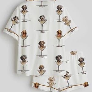 Op de afbeelding: Witte pyjamaset met een herhaald patroon van teckels in martiniglazen. De top met korte mouwen en de shorts hebben bruine biezen. De teckels zijn in verschillende kleuren, waaronder bruin, zwart en bruin.