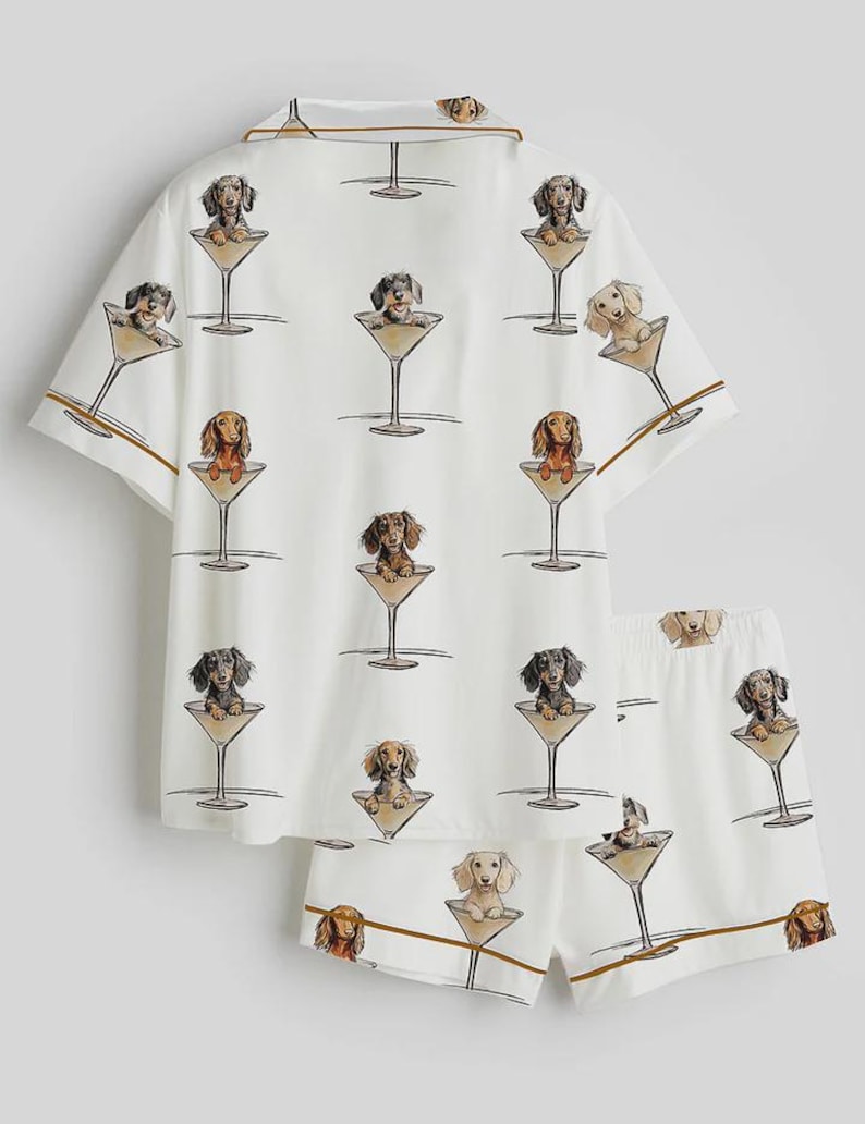 Op de afbeelding: Witte pyjamaset met een herhaald patroon van teckels in martiniglazen. De top met korte mouwen en de shorts hebben bruine biezen. De teckels zijn in verschillende kleuren, waaronder bruin, zwart en bruin.