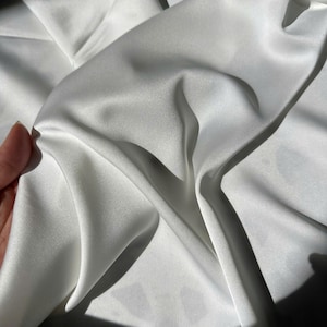 K&ouml;nnte beinhalten: Ein St&uuml;ck wei&szlig;er Stoff mit einer glatten, gl&auml;nzenden und satinartigen Oberfl&auml;che. Das Material ist in weichen, flie&szlig;enden Falten drapiert, die tiefe Schatten und helle Glanzlichter auf der Oberfl&auml;che erzeugen und die Reflexion des Textils betonen.