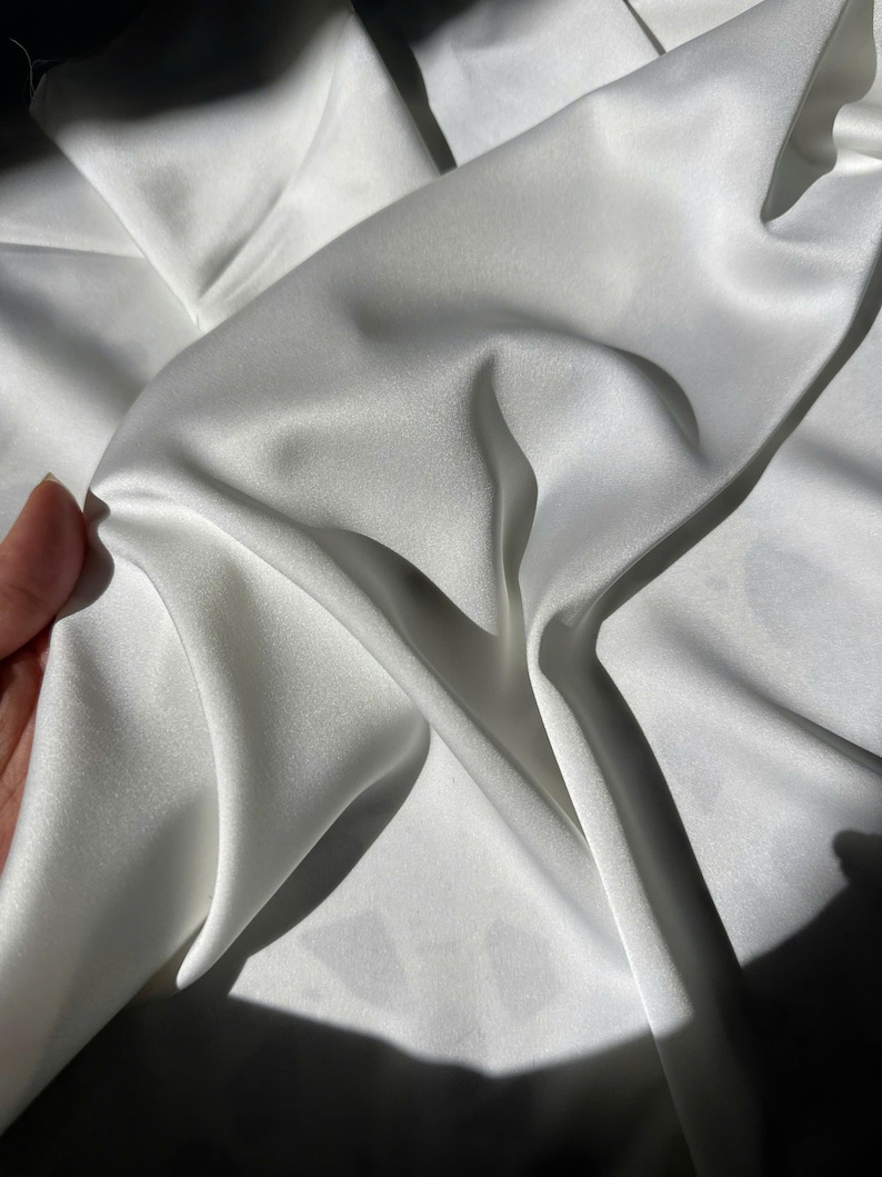 K&ouml;nnte beinhalten: Ein St&uuml;ck wei&szlig;er Stoff mit einer glatten, gl&auml;nzenden und satinartigen Oberfl&auml;che. Das Material ist in weichen, flie&szlig;enden Falten drapiert, die tiefe Schatten und helle Glanzlichter auf der Oberfl&auml;che erzeugen und die Reflexion des Textils betonen.