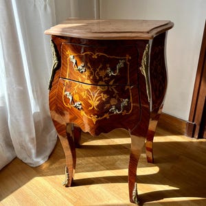 May include: A two-drawer antique wooden bedside table with a curved front. The table has a light-coloured top and is decorated with floral marquetry and brass hardware. The wood has a glossy finish and is set on curved legs.