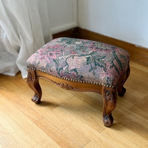 May include: A rectangular footstool featuring a wooden frame with carved cabriole legs and a cushioned top upholstered in a floral tapestry fabric. The fabric displays a pattern of pink, green, and grey flowers, accented by a row of brass nailhead trim.