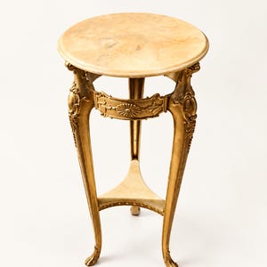 May include: A round side table featuring a light beige marble top and a gold-finished metal base. The three curved legs are decorated with ornate carvings and connected by a triangular lower shelf with a matching marble surface.