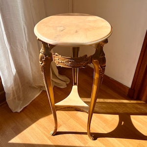 Peut inclure: Un petit support de plante de style antique avec un dessus rond en marbre clair. Le support présente des pieds dorés ornés et des sculptures décoratives. Le support mesure environ 71 cm de haut.