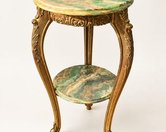 May include: A round side table featuring a green and brown marbled stone top and a matching lower shelf. The table has three curved, gold-toned legs with ornate carved detailing along the frame and feet, providing a sturdy structure.