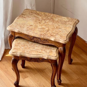 Puede incluir: Un juego de dos mesas nido con tableros rectangulares de patrón marmolado en tonos beige y marrón. Las mesas tienen estructuras de madera marrón oscuro con patas cabriolé curvas y detalles decorativos tallados en forma de concha.