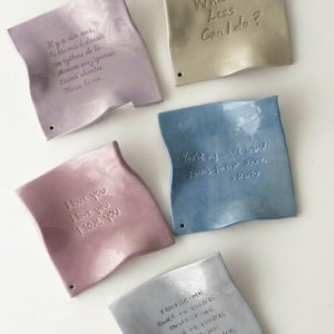 Puede incluir: Cinco platos de cerámica con forma de nota en colores pastel. Cada uno tiene un mensaje manuscrito único, incluyendo frases como "Te amo" y "Eres mi mundo entero". Los platos tienen un aspecto ligeramente arrugado y pequeños agujeros.