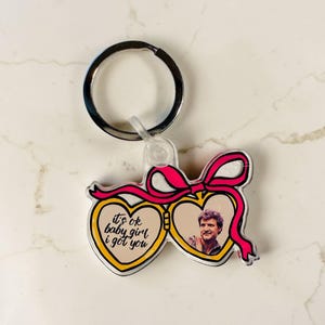 May include: A heart-shaped keychain with a pink bow and a silver key ring. One heart has the text "it's ok baby girl i got you" in black. The other heart contains a photo of a man.