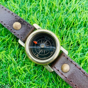 May include: A vintage compass watch with a brown leather strap. The compass has a gold-coloured bezel and a black face with directional markings. The strap is secured with brass-coloured rivets, set against a backdrop of green grass.