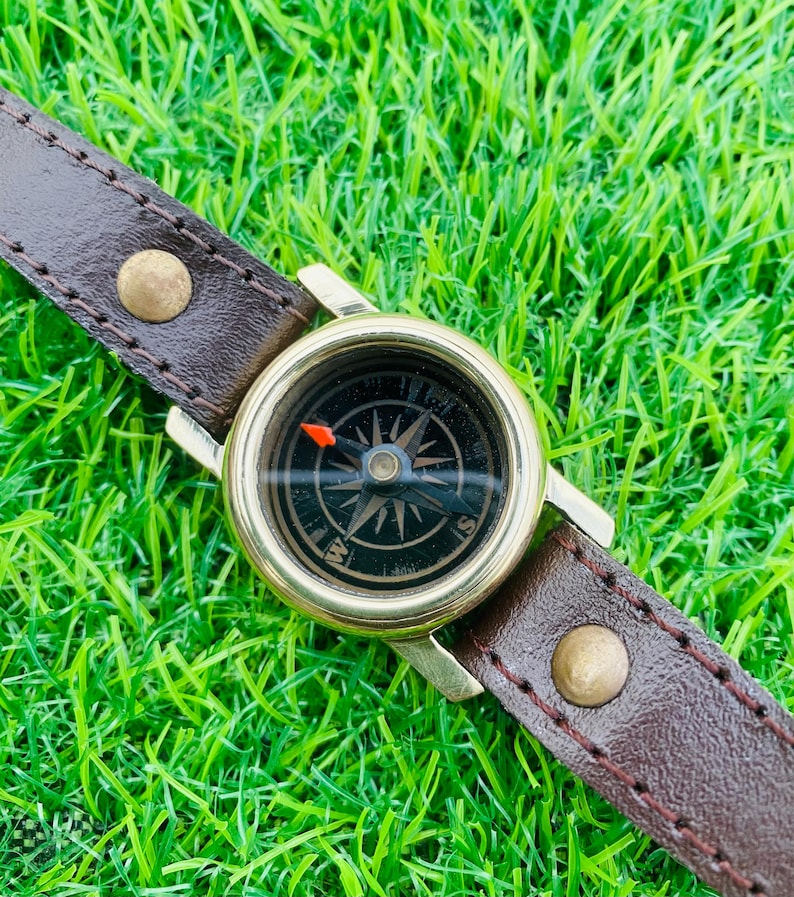May include: A vintage compass watch with a brown leather strap. The compass has a gold-coloured bezel and a black face with directional markings. The strap is secured with brass-coloured rivets, set against a backdrop of green grass.
