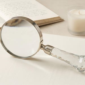 May include: A magnifying glass featuring a circular silver-toned metal frame and a clear, faceted crystal handle. The lens is transparent, and the handle has a geometric, multi-sided cut design that reflects light, attached to the frame by a silver metal collar.
