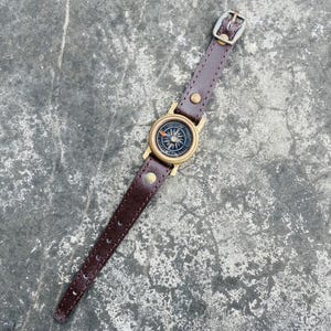 May include: A brown leather compass watch with a brass compass face. The watch has a dark brown leather strap with a buckle. The compass face features directional markings and a needle. The watch is designed for navigation and outdoor activities.