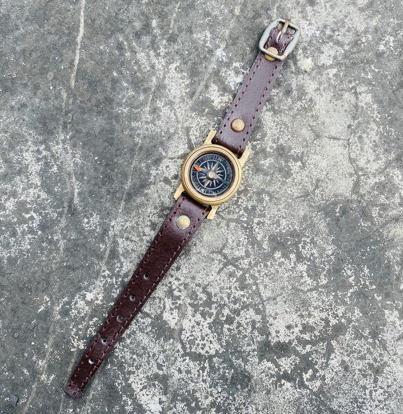 May include: A brown leather compass watch with a brass compass face. The watch has a dark brown leather strap with a buckle. The compass face features directional markings and a needle. The watch is designed for navigation and outdoor activities.