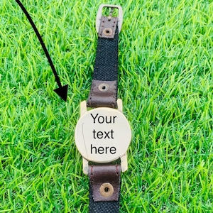 May include: A wristwatch with a black fabric strap and brown leather detailing. The gold-coloured watch face displays the text "Your text here". The watch is set against a background of green grass.