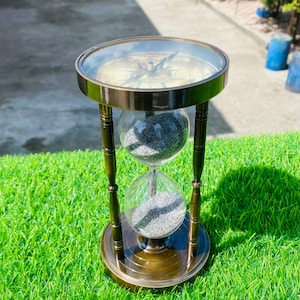 May include: An antique brass hourglass with a compass on top. The hourglass has two glass bulbs connected by a narrow neck, filled with gray sand. The compass features directional markings. The hourglass is supported by a brass frame with decorative legs, and sits on a round base.