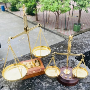 May include: Two brass balance scales with gold-coloured pans and chains. One scale sits on a wooden base with star-shaped accents, and the other on a dark brown base. The scales are designed with a classic, vintage aesthetic.