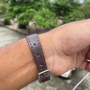 May include: A close-up of a brown leather watch strap with a silver buckle. The strap has visible stitching and is attached to a wrist. The background is blurred, with green foliage and a glimpse of a building.