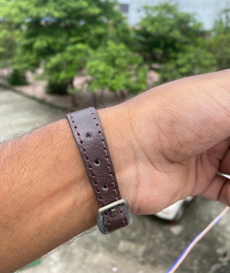 May include: A close-up of a brown leather watch strap with a silver buckle. The strap has visible stitching and is attached to a wrist. The background is blurred, with green foliage and a glimpse of a building.