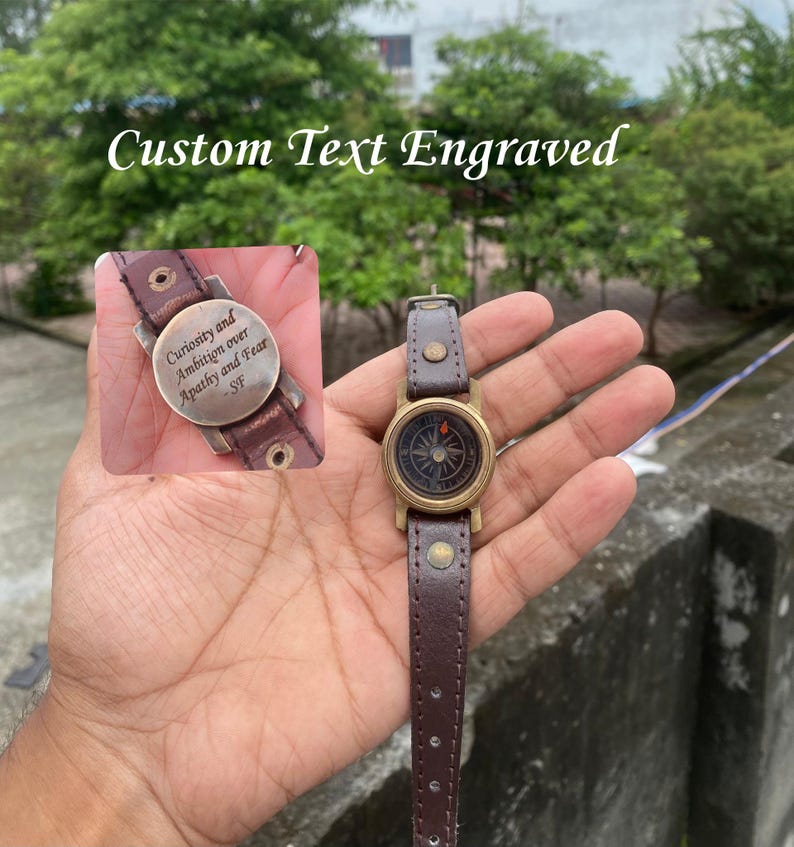May include: A brown leather compass watch with a brass bezel and a dark face. The watch strap is brown leather. The image also shows a close-up of the watch's engraved plate with the text "Curiosity and Ambition over Apathy and Fear - SF". The text "Custom Text Engraved" is at the top.