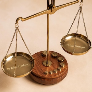 Puede incluir: Una balanza de latón con una base circular de madera que incluye varios pesos pequeños de latón. El plato izquierdo tiene grabado el texto 10 Jahre Apotheke y el plato derecho tiene grabado el texto Urlich Terstegen. La balanza tiene dos platos colgantes.