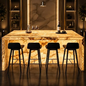 May include: A large, rectangular, backlit onyx kitchen island with a warm, glowing amber and brown veined pattern. Four black bar stools with curved backrests are positioned in front of the island. A glass decanter, two wine glasses, and a wine bottle are placed on the surface.
