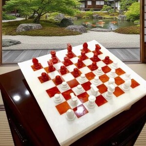 Könnte beinhalten: Ein Schachspiel mit roten und weißen Figuren auf einem weiß-orangen Schachbrett. Das Brett steht auf einem dunkelbraunen Tisch. Der Hintergrund zeigt einen japanischen Garten mit einem Teich und Bäumen.