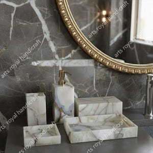 Könnte beinhalten: Ein Badezimmer-Set aus weißem Marmor mit grauer Maserung. Das Set umfasst einen Seifenspender, einen Zahnbürstenhalter, ein kleines Tablett, eine rechteckige Box und ein größeres Tablett. Im Hintergrund befindet sich ein goldgerahmter Spiegel.