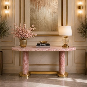 Op de afbeelding: Een consoletafel met een blad van gepolijst rozenkwarts en twee cilindrische rozenkwarts poten verbonden door een gouden metalen voet. De tafel bevat een gouden vaas met roze bloemen, een gouden schaal, twee gestapelde boeken met Chanel en Dior, en een lamp.
