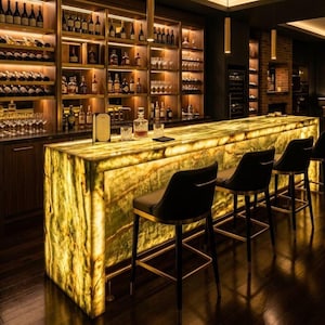 May include: A backlit green onyx bar counter with a polished stone surface, featuring a warm internal glow. Four black upholstered bar stools with gold-toned metal frames are positioned along the front, set against a dark wood bar back with illuminated shelving filled with various glass bottles and stemware.