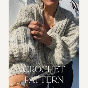 Op de afbeelding: Een lichtgrijs gehaakt vest met zwarte vlekken, met een geribbeld ontwerp en volumineuze mouwen. De tekst "CROCHET PATTERN" wordt in het wit weergegeven. Een gouden armband en ketting zijn ook zichtbaar.