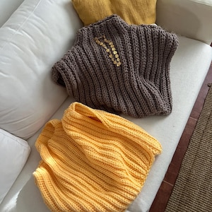 Op de afbeelding: Twee handgemaakte gehaakte tops. Eén is een mosterdgele col, de andere is een bruine, geribbelde mouwloze top met gouden versieringen. Beide worden gepresenteerd op een witte bank met een geel kussen.