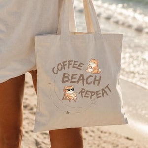 Könnte beinhalten: Beige Tragetasche mit dem Aufdruck "COFFEE BEACH REPEAT". Die Tasche zeigt einen Cartoon Shiba Inu-Hund mit Sonnenbrille und Strandstuhl. Die Person trägt ein weißes T-Shirt und steht an einem Sandstrand.